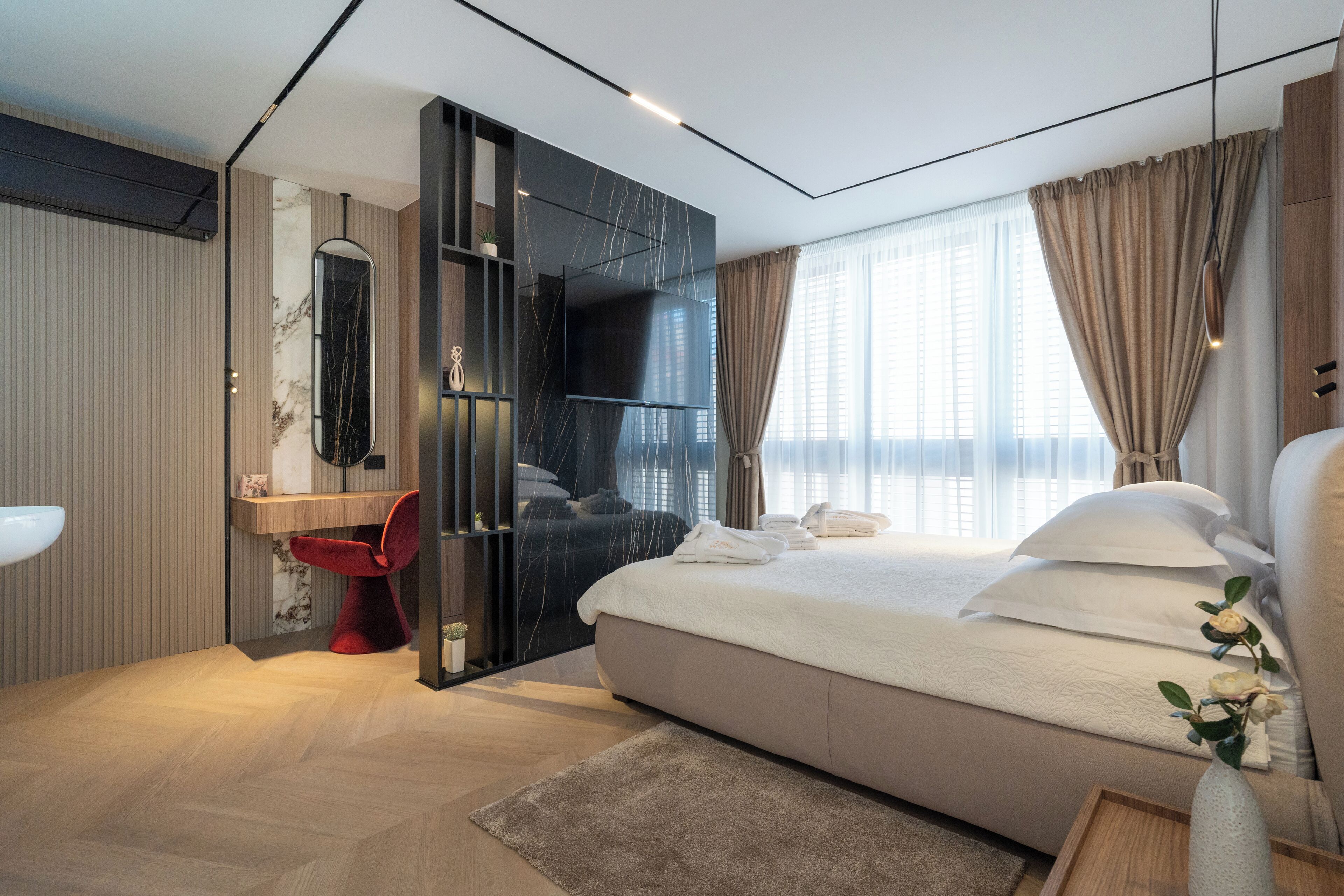 A modern bedroom with a large bed, a built-in TV, a vanity with a mirror, and a red chair.