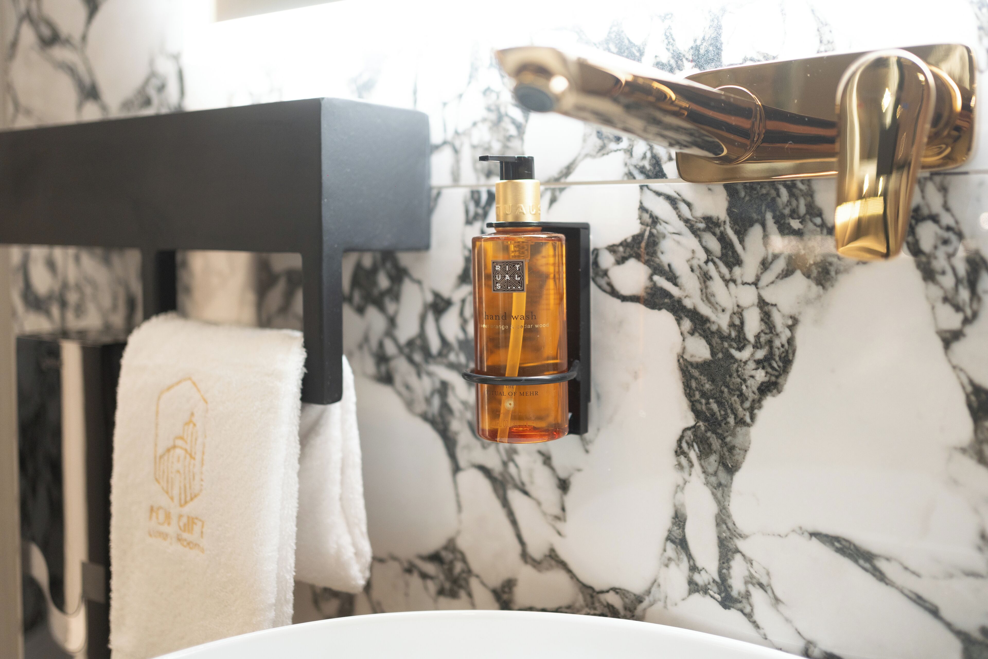 Un baño con pared de mármol, grifo dorado, toallero negro y una botella de jabón de manos.