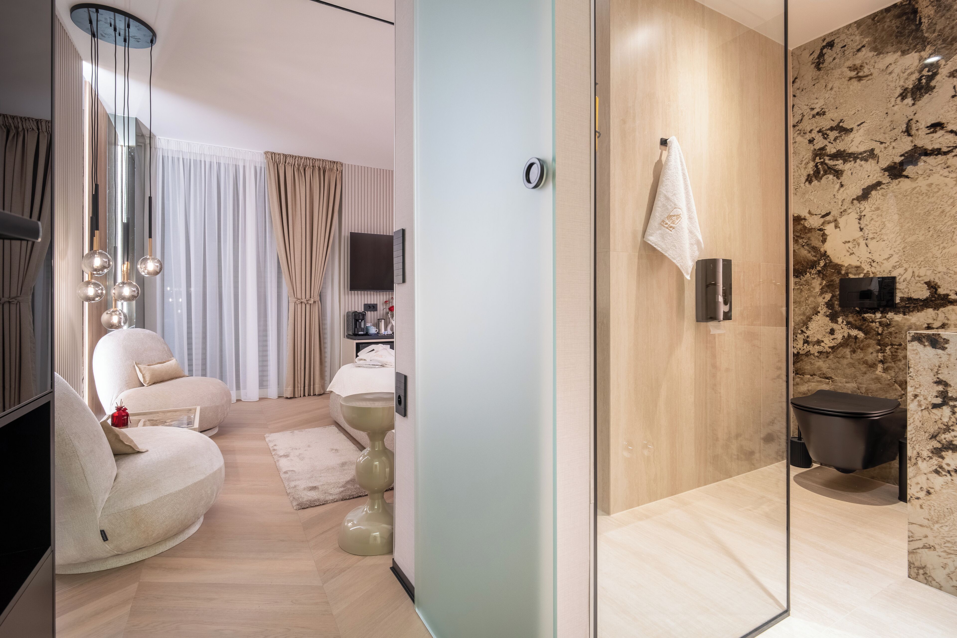 Quarto de hotel moderno com divisória de vidro, sofá, televisão, TV de tela plana, cama, mesa de cabeceira e porta-toalhas fixado na parede.