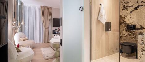 A modern hotel room with a glass partition, a sofa, a TV, a flat-screen TV, a bed, a bedside table, and a wall-mounted towel rack.