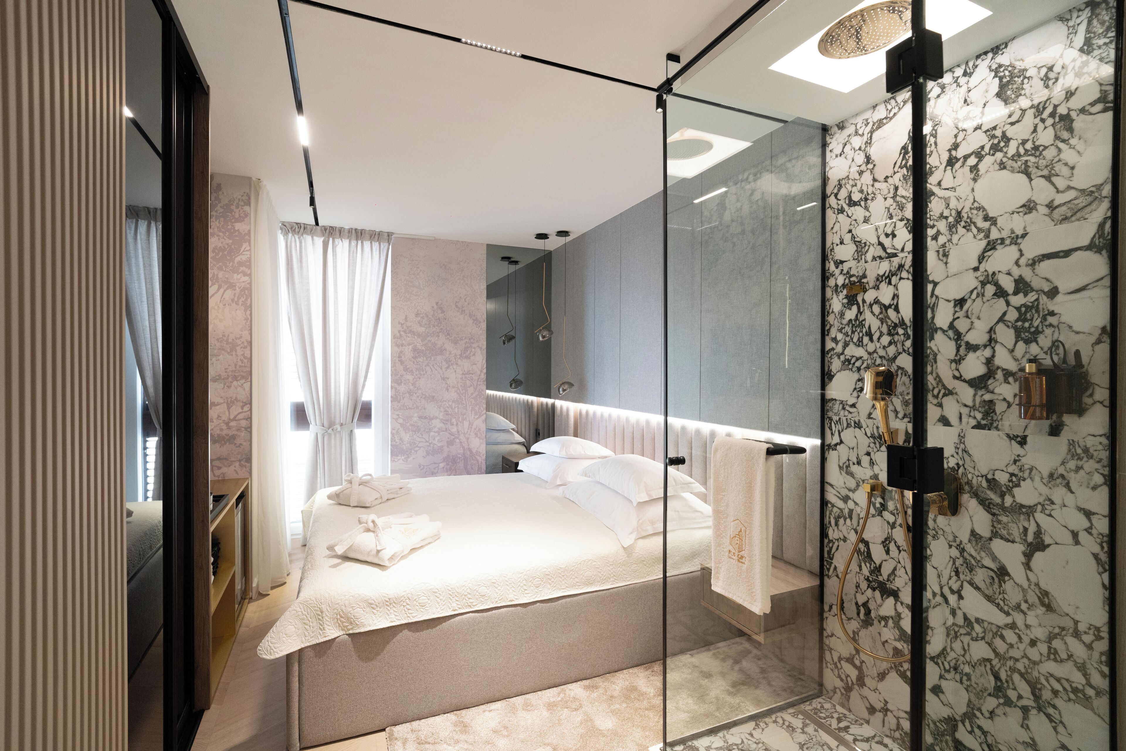 A modern bedroom with a glass shower enclosure, a bed with white linens, and a marble-patterned wall.