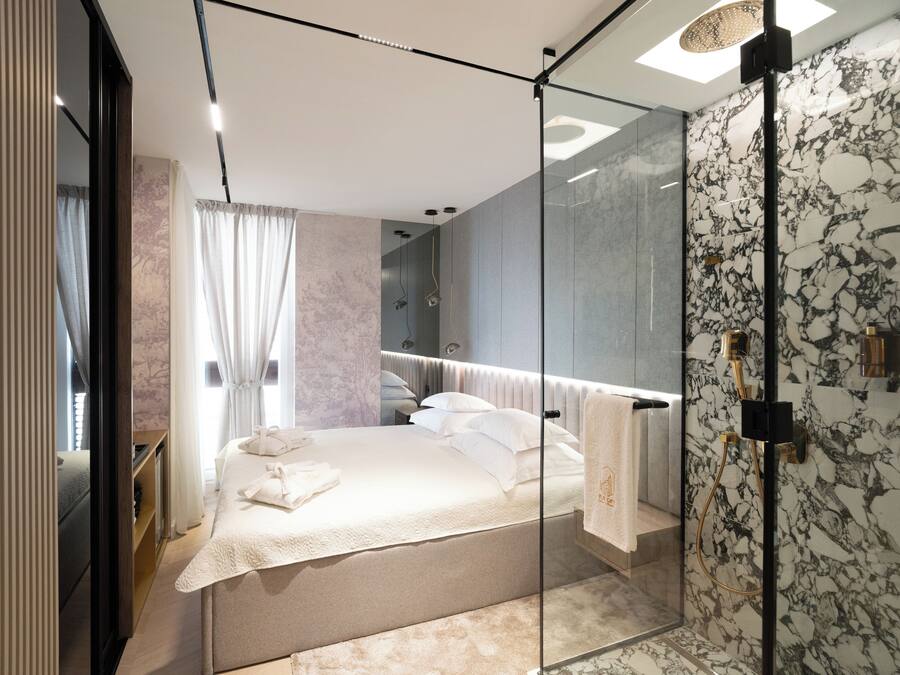 A modern bedroom with a glass shower enclosure, a bed with white linens, and a marble-patterned wall.