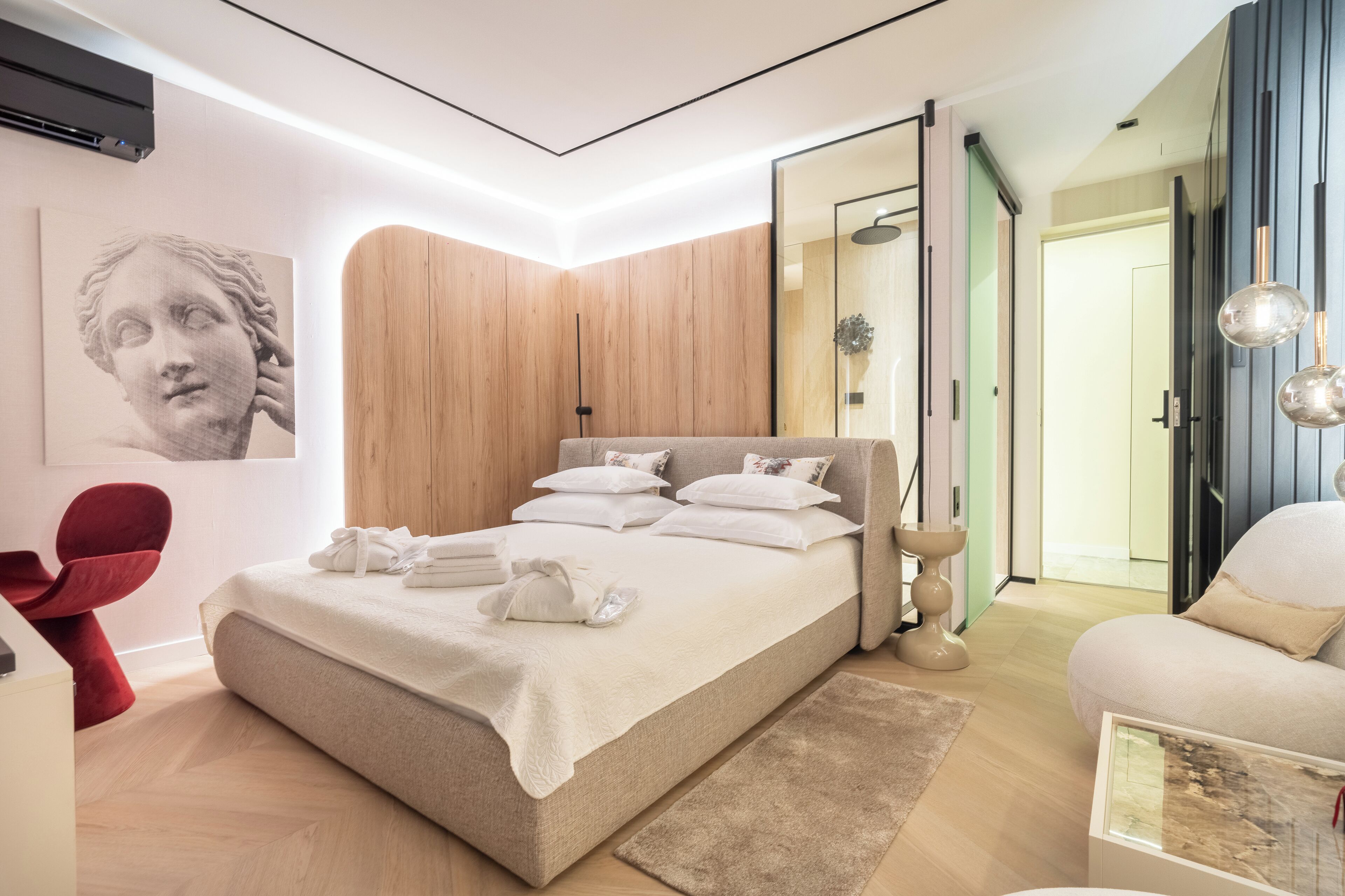 A modern bedroom with a large bed, wooden panel walls, a red chair, and a glass table.