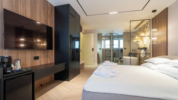 Una habitación de hotel moderna con una cama grande, un televisor de pantalla plana montado en la pared, una kitchenette pequeña y un vestidor.