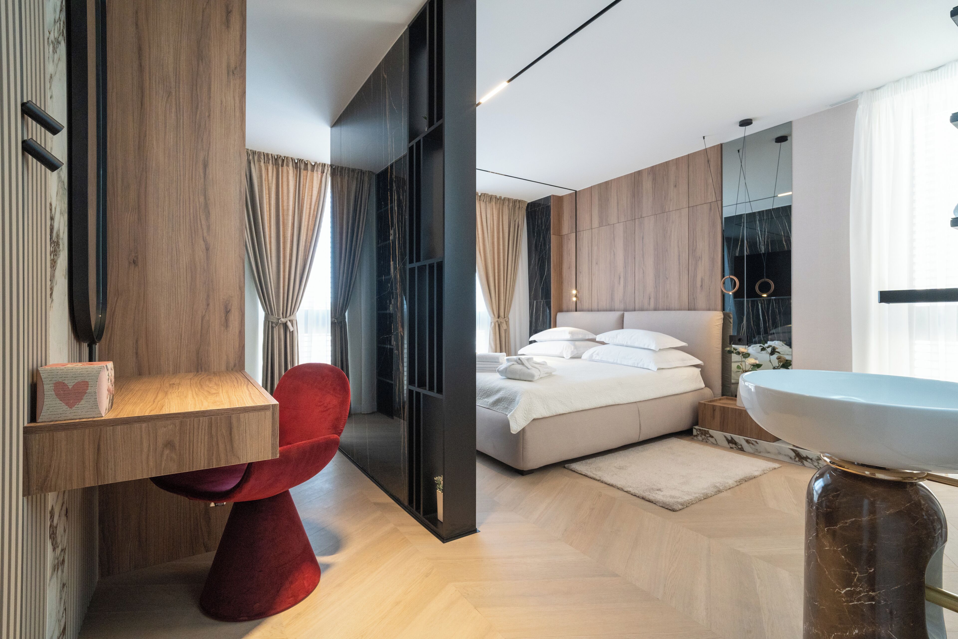 A modern bedroom with a bed, a red chair, a wooden desk, and a bathroom with a sink and mirror.