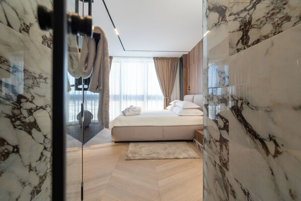 A modern bedroom with a large bed, a wooden floor, and a glass door leading to a bathroom. - Luxury rooms FOR GIFT (Zadar)