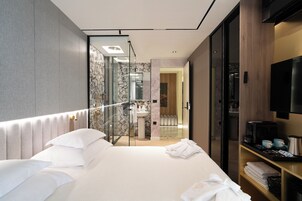 Quarto de hotel moderno com uma cama grande, casa de banho com paredes de vidro, roupeiro de madeira e televisão de ecrã plano.