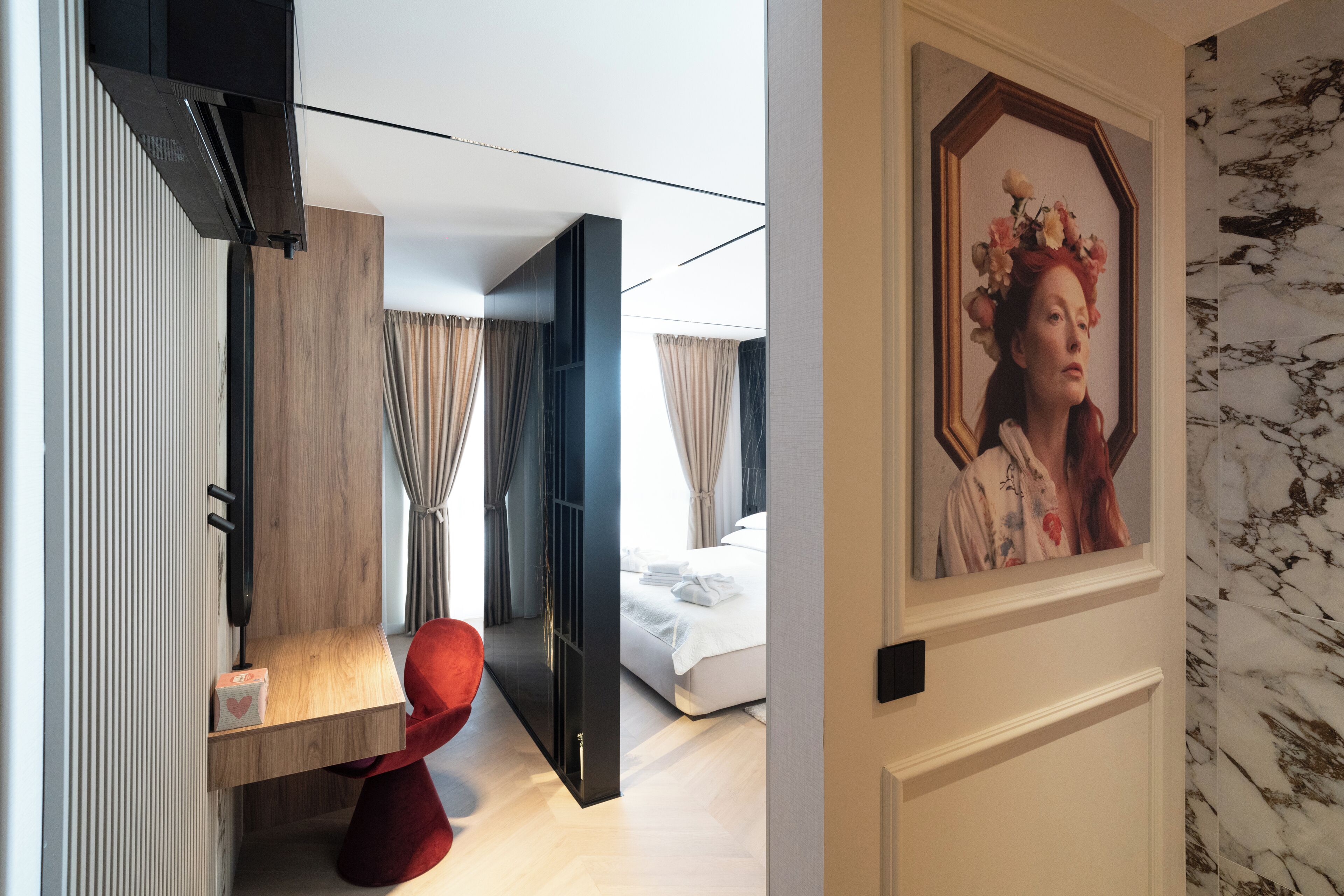 A modern hotel room with a wooden floor, a red chair, a bed with white linens, and a framed picture on the wall.