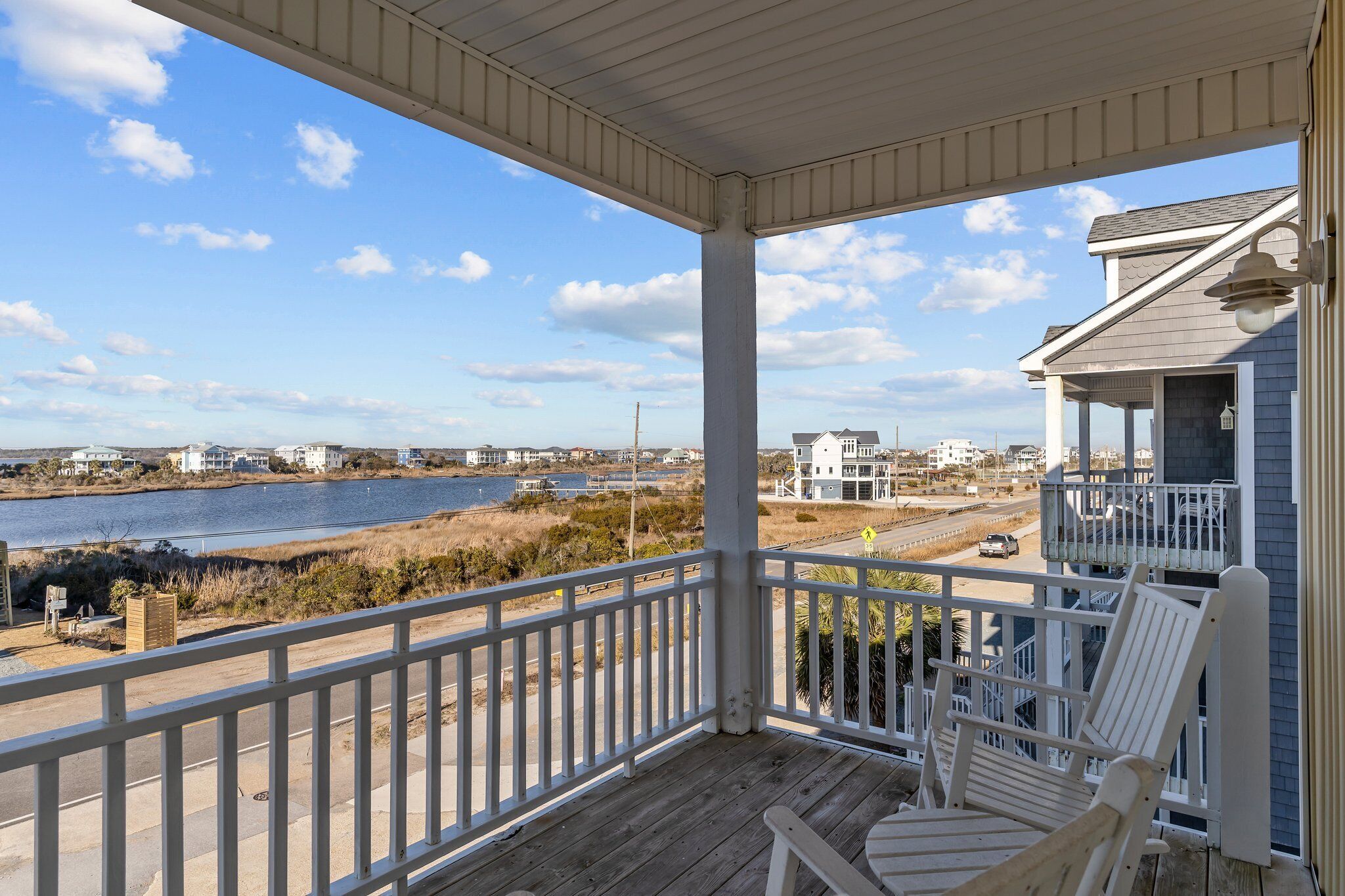 Eine überdachte Veranda mit weißen Geländern, einer Holzterrasse und Blick auf ein Gewässer sowie Strandhäuser in der Ferne.