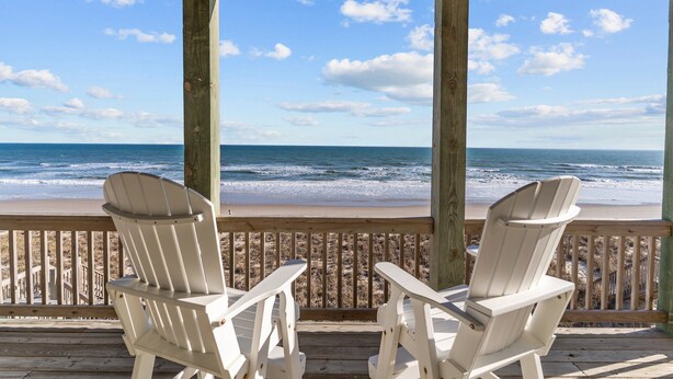 A wooden deck with two white Adirondack chairs facing a beach.