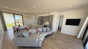 A modern living room with a grey sectional sofa, a dining area with a table and chairs, a kitchen area, and a large TV mounted on the wall. - Zion on Yorkes-2 bedroom downstairs unit - Off peak special! (Edithburgh)
