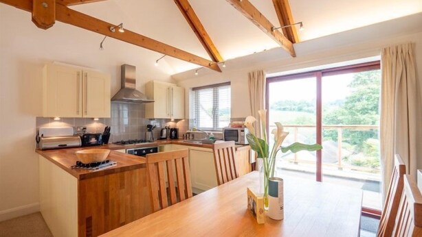 A kitchen with wooden beams, a dining area with a table and chairs, and a view of greenery outside.