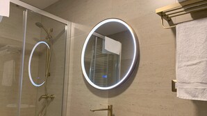 A modern bathroom with a glass-enclosed shower, a round mirror with LED lighting, a wooden vanity with a white sink, and toiletries. - Cumbres de Chiloé (Castro)
