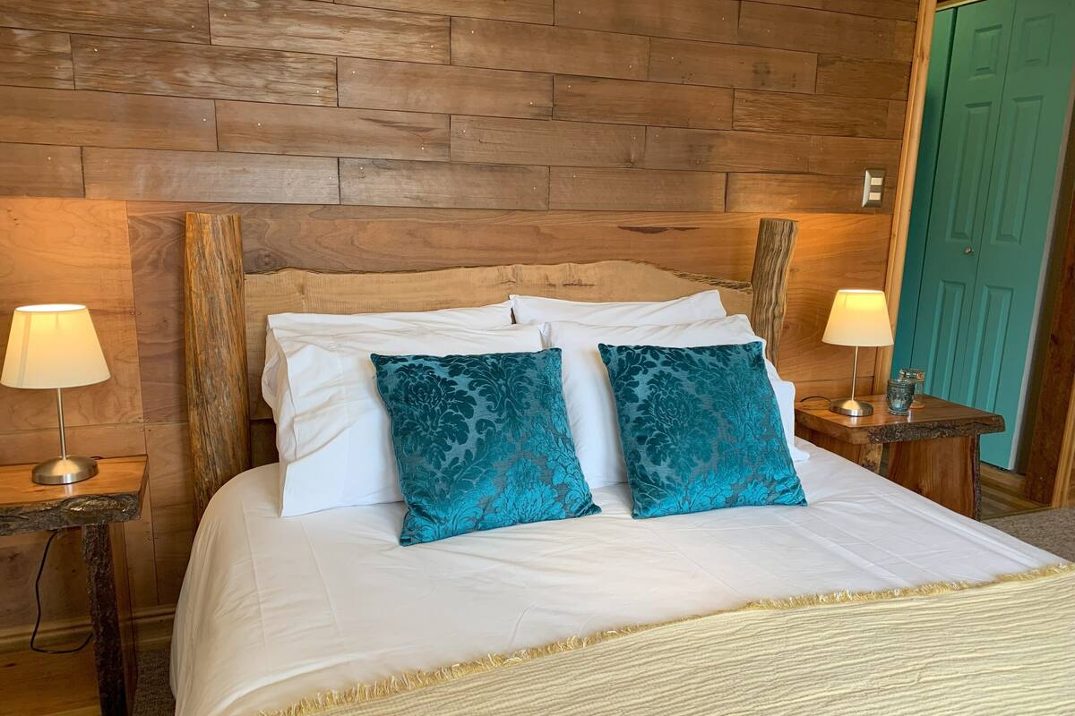 Ein ordentlich bezogenes Bett mit weißen Leinentüchern und zwei Kissen in Türkis-Muster, eingerahmt von Nachttischen aus Holz mit Lampen, vor einer Holzwand.