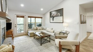 A spacious living room with a sofa, armchair, coffee table, and a television. There is a large window with a view of trees, a fireplace, and a framed picture on the wall. - River Pines A303 3 Bedroom Condo (Edwards)