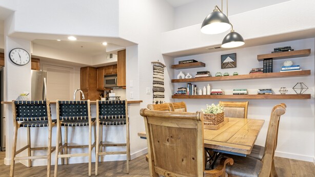 A dining area with a wooden table and chairs, a clock on the wall, and shelves with books and decorative items. - River Pines A303 3 Bedroom Condo (Edwards)