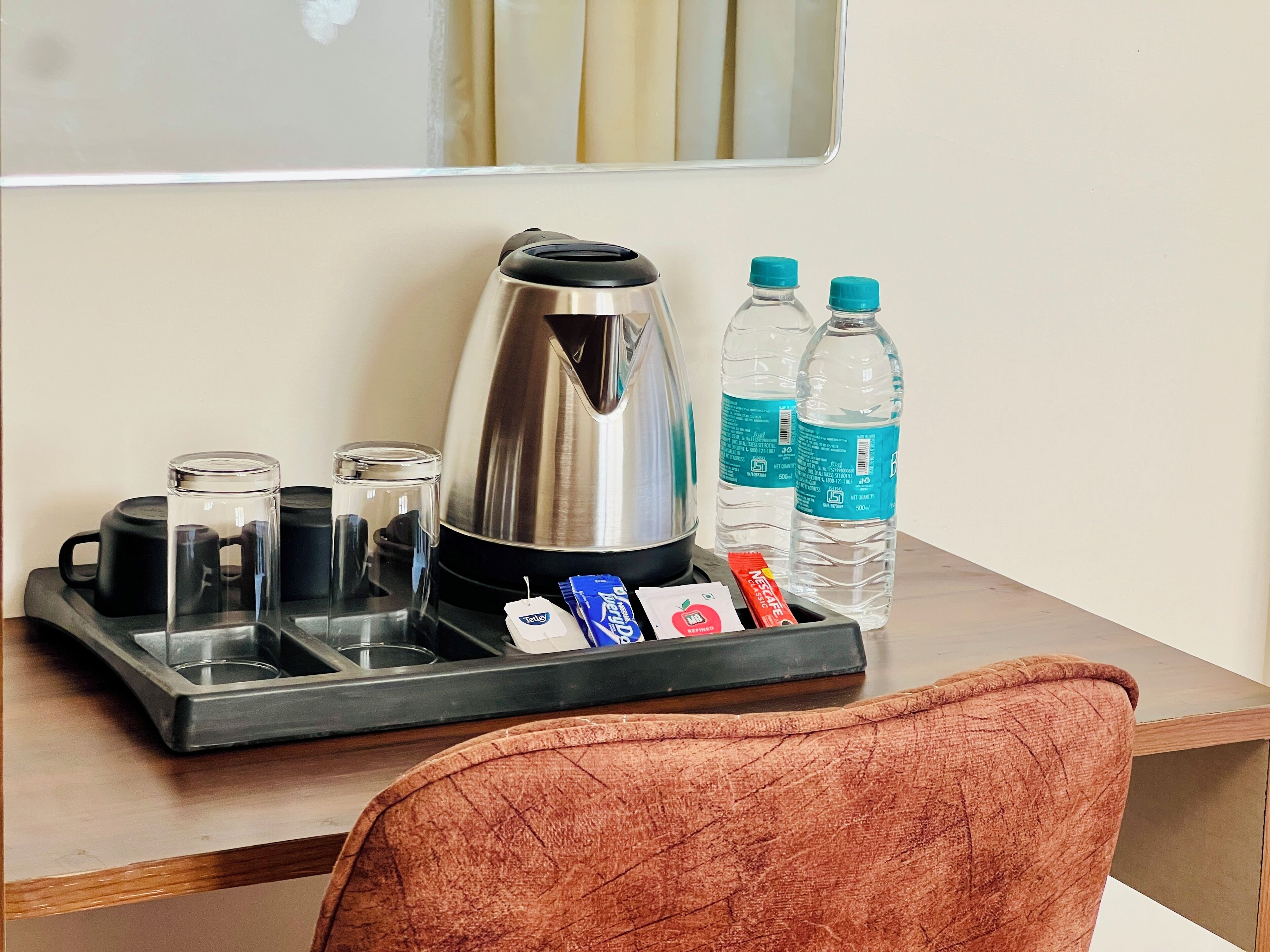Camera d'albergo con una macchina per il caffè, due bottiglie d'acqua, un vassoio con tazze e una bustina di tè, e una sedia.