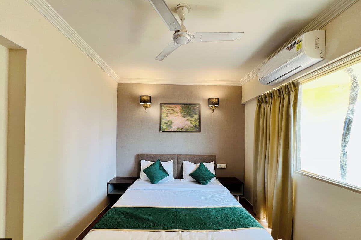 A hotel room with a bed, a ceiling fan, wall-mounted lights, a window with curtains, and an air conditioning unit.