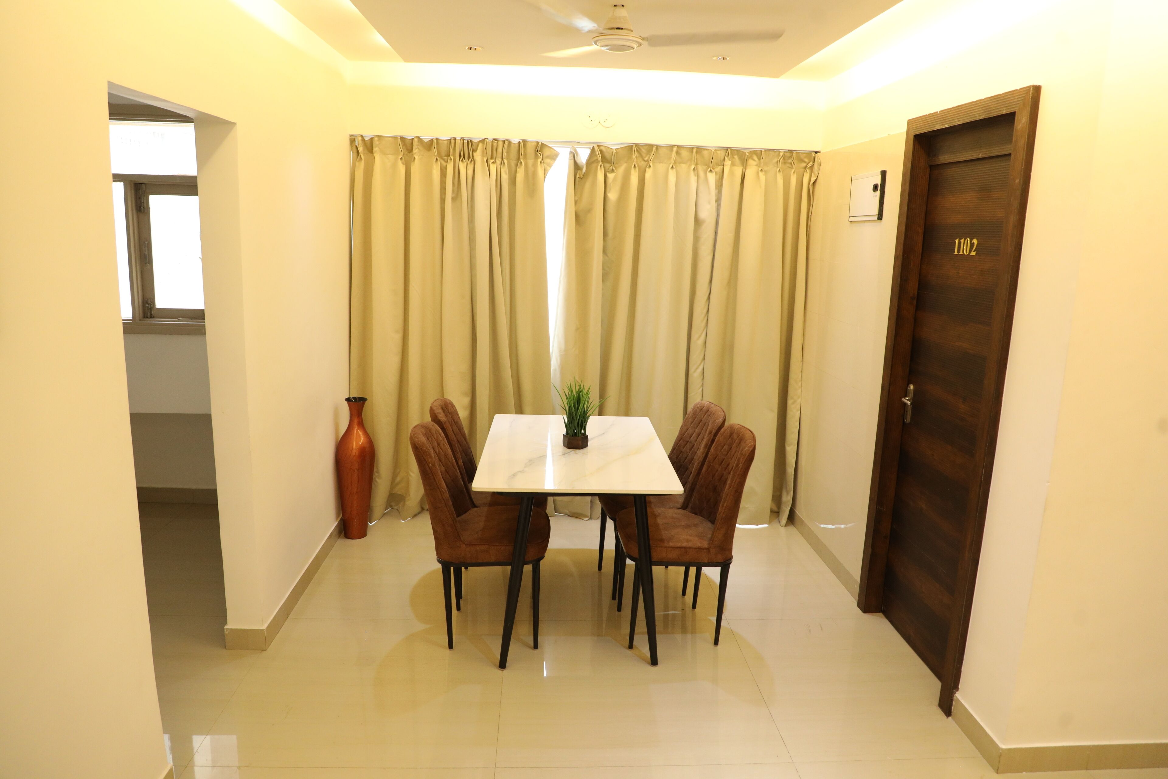 A dining area with a table and four chairs, a potted plant, and a door with the number 1102.