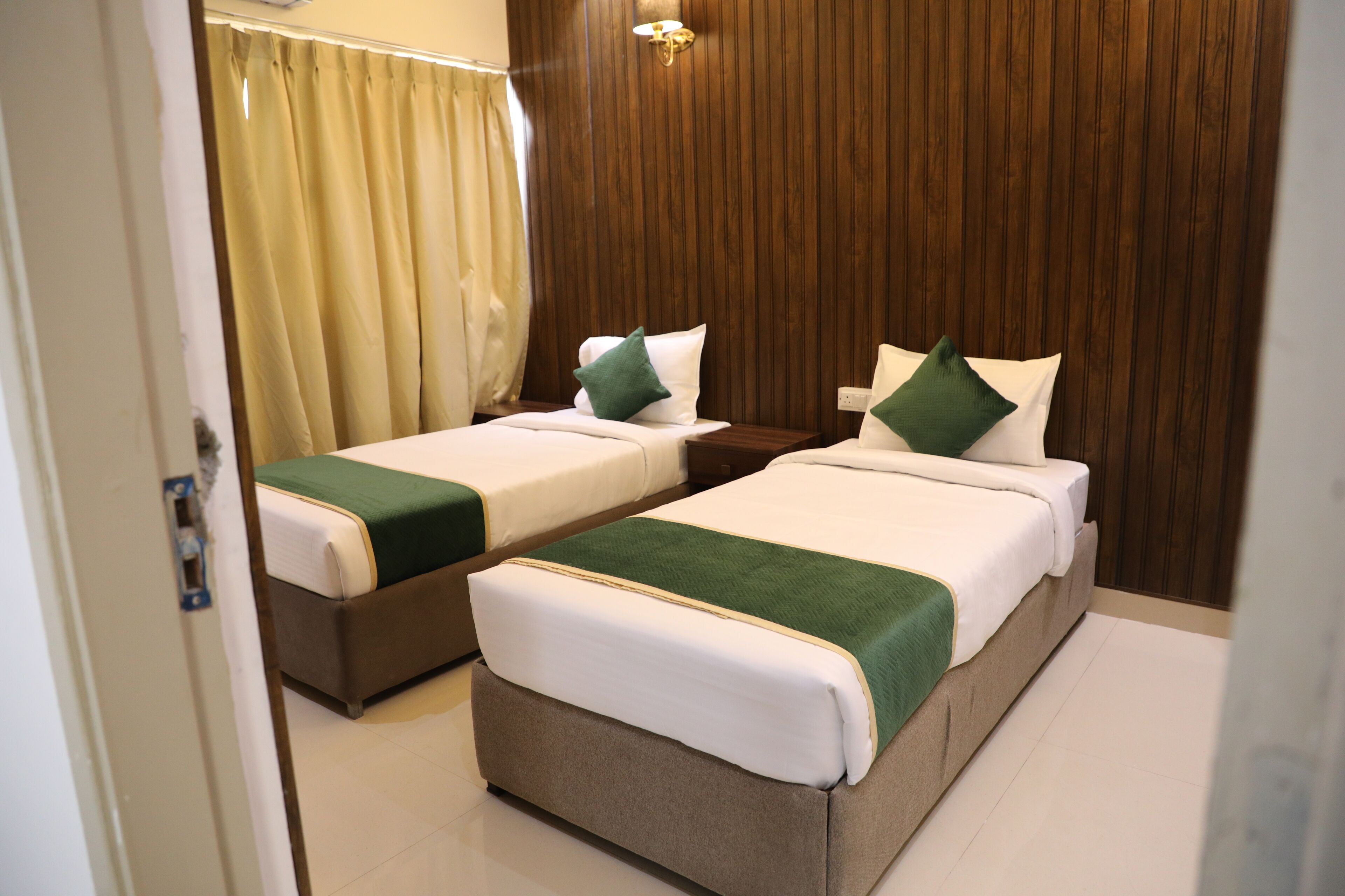 A hotel room with two beds, each with a green and white bedspread, wooden headboard, and a nightstand with a lamp.