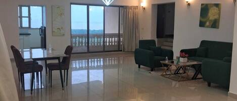 A spacious living area with a dining table, two green sofas, and a coffee table. There are large windows with a view of the outdoors.