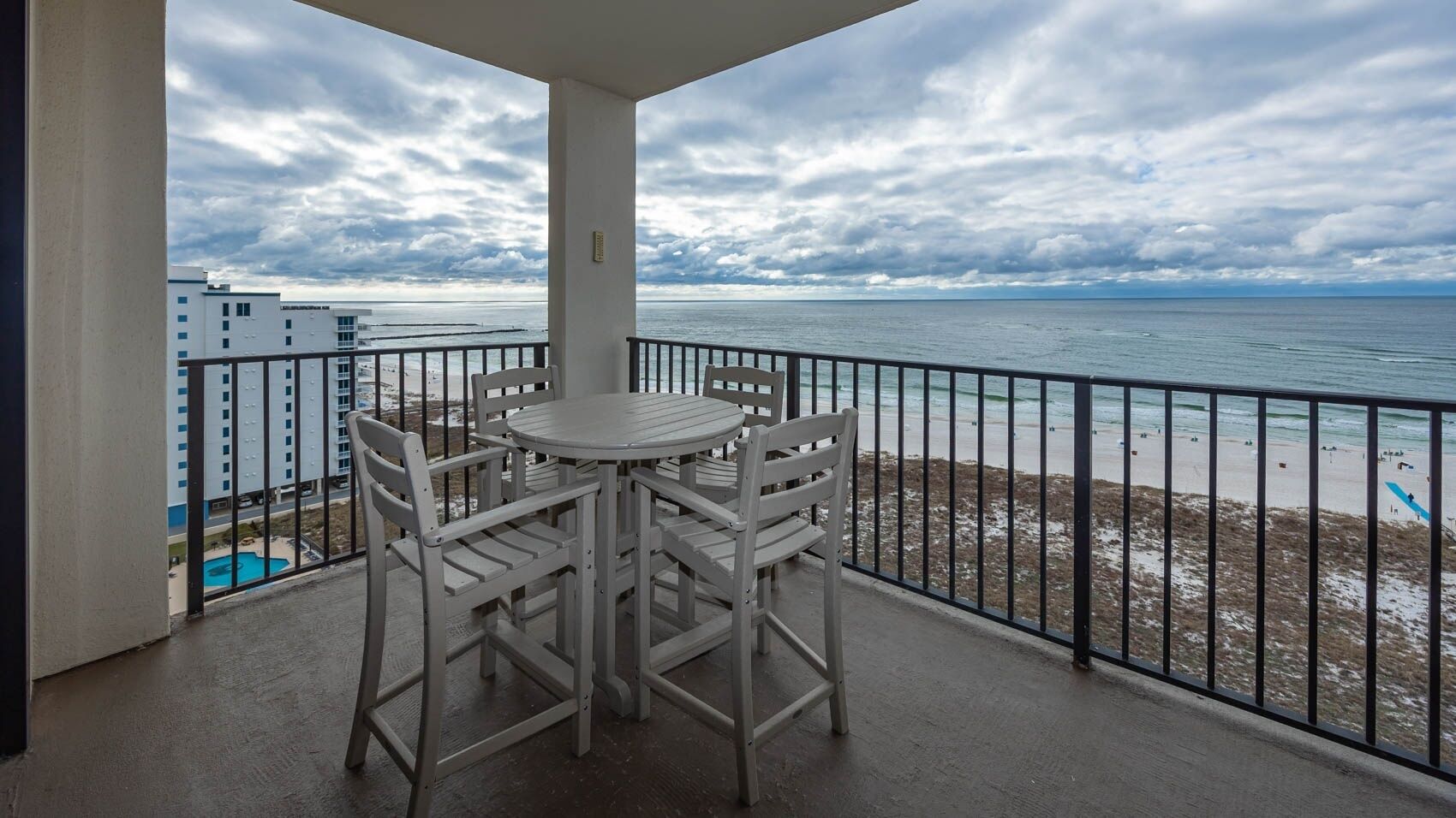 A balcony with a round table and four chairs, overlooking a beach with a view of the ocean and a cloudy sky.