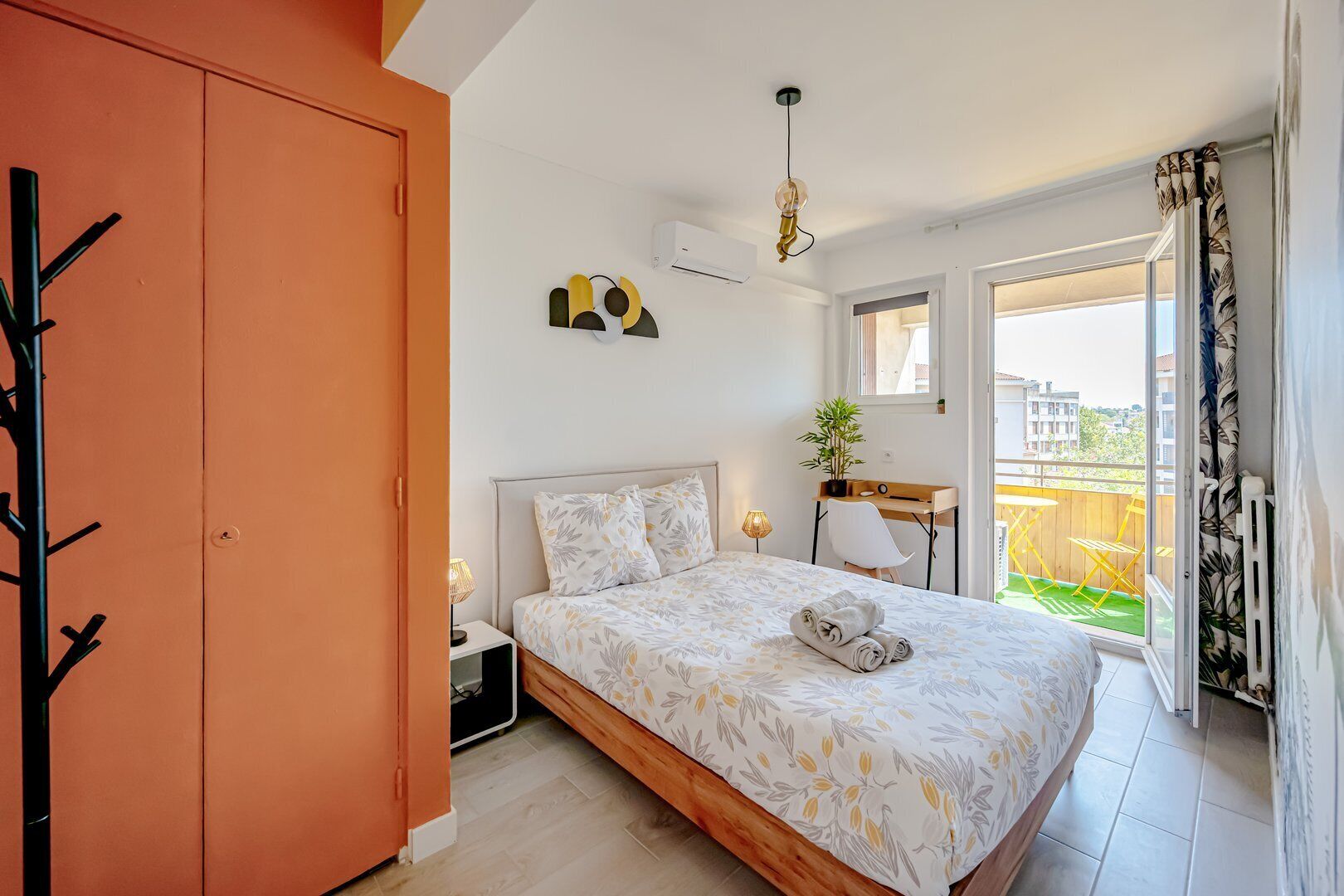 Ein Schlafzimmer mit Bett, Schreibtisch, Stuhl, einer Pflanze und Blick ins Freie.
