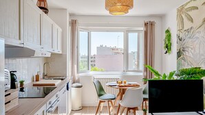 A modern kitchen with white cabinets, a dining area with a table and chairs, a large window with a view of buildings, and a flat-screen TV on a stand. - Wisdom Monkeys Apartment - Balcony & AC (Toulon)