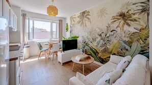 A modern living room with a large tropical mural, a dining area with a white table and chairs, and a kitchen with white cabinets and a large window. - Wisdom Monkeys Apartment - Balcony & AC (Toulon)