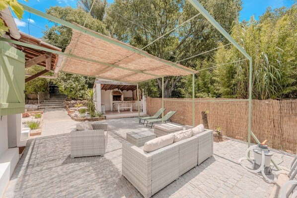 Eine überdachte Terrasse im Freien mit Korbmöbeln, einem Grillbereich und einem gepflasterten Weg, der zu einer Treppe führt.