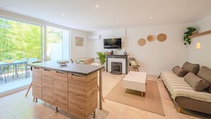 Un salon moderne avec un canapé, un coin repas comprenant une table et des chaises, une cuisine équipée d’une télévision et d’une cheminée, et une grande fenêtre donnant sur un paysage verdoyant.