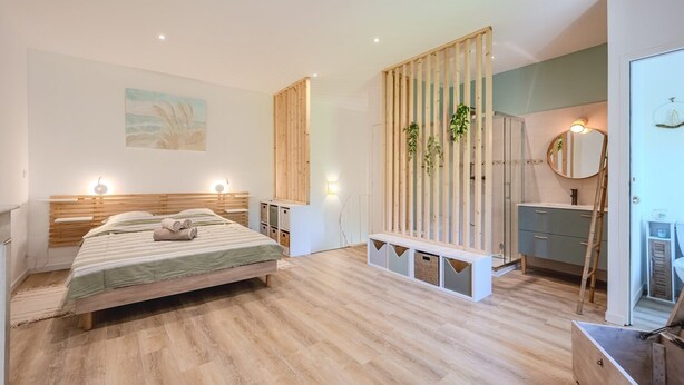 Une chambre moderne avec un lit en bois, des tables de chevet, un banc intégré avec espace de rangement, une salle de bain avec un miroir et un lavabo, et une cloison en bois agrémentée de plantes.