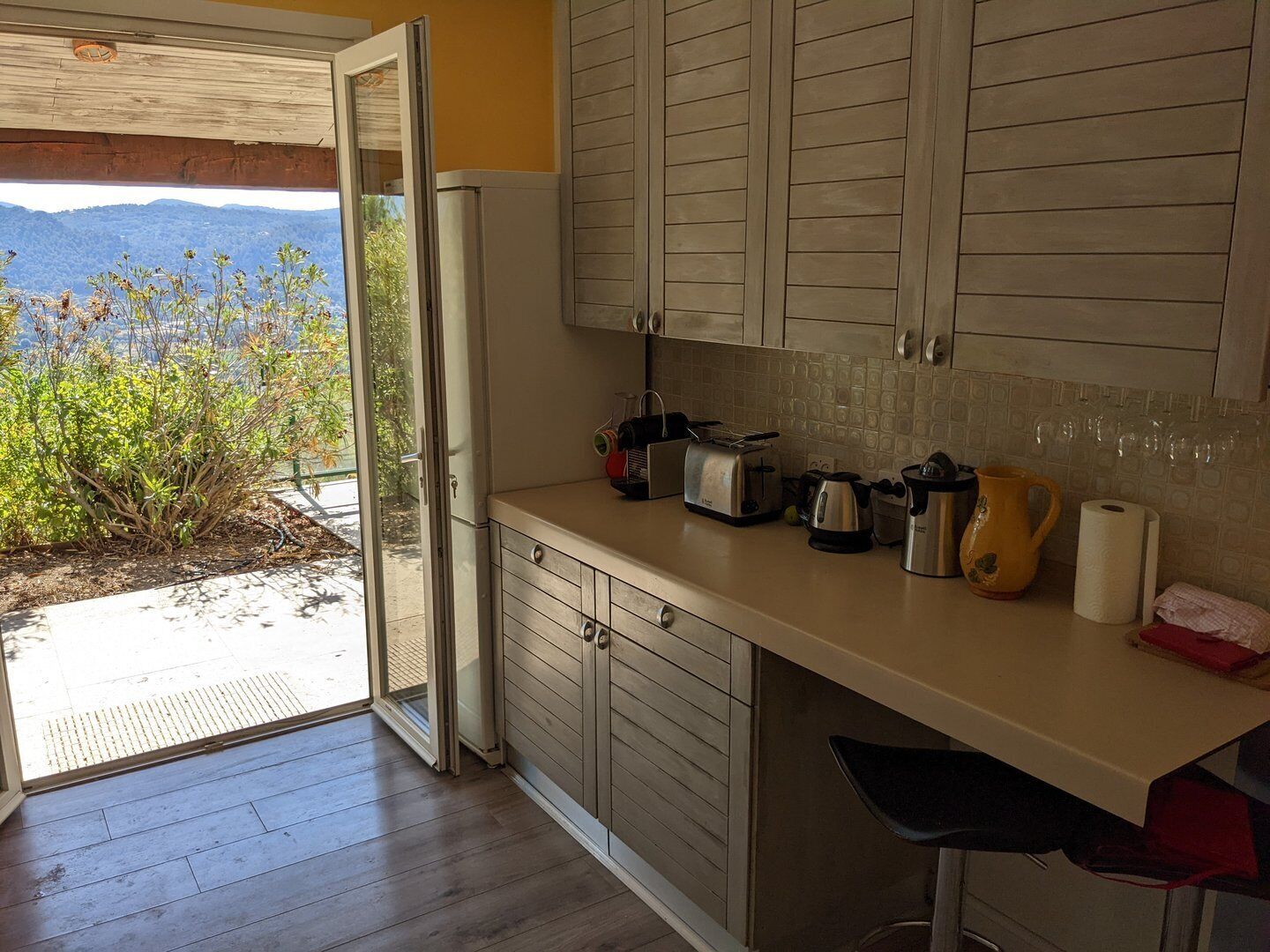 A kitchen with wooden cabinets, a countertop with a toaster, kettle, and coffee pot, and a view of mountains through an open door.