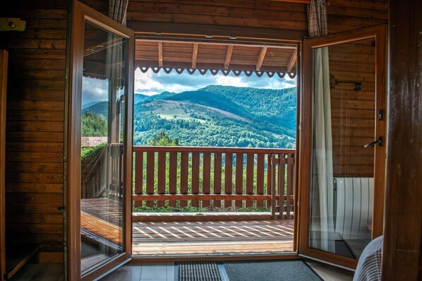 A wooden cabin with an open door revealing a balcony with a view of a forested mountain range. - Entire chalet, panoramic views, stream and sauna. (Bussang)