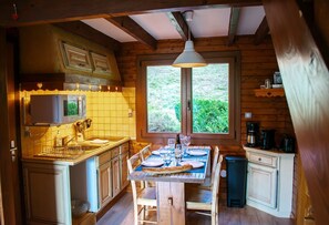 A cozy kitchen with wooden cabinets, a dining table set for two, and a window overlooking greenery. - Entire chalet, panoramic views, stream and sauna. (Bussang)