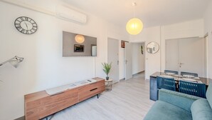Una sala moderna con una consola de madera, un reloj redondo, un televisor montado y un área de comedor con sillas azules.