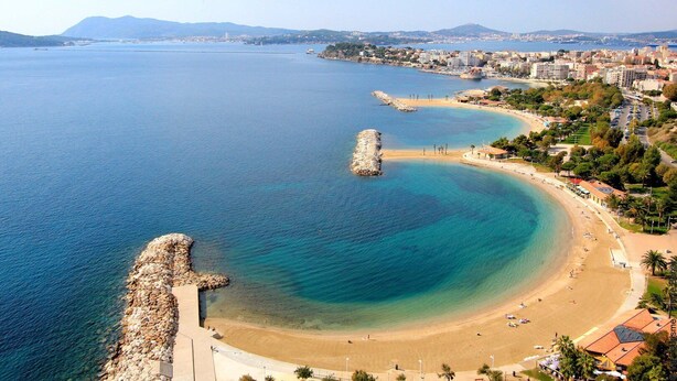 Un paisaje urbano costero con playa, aguas azules cristalinas y un rompeolas de piedra.