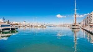 Un puerto deportivo con varias embarcaciones amarradas, destacándose un velero, y un edificio con balcones al fondo.