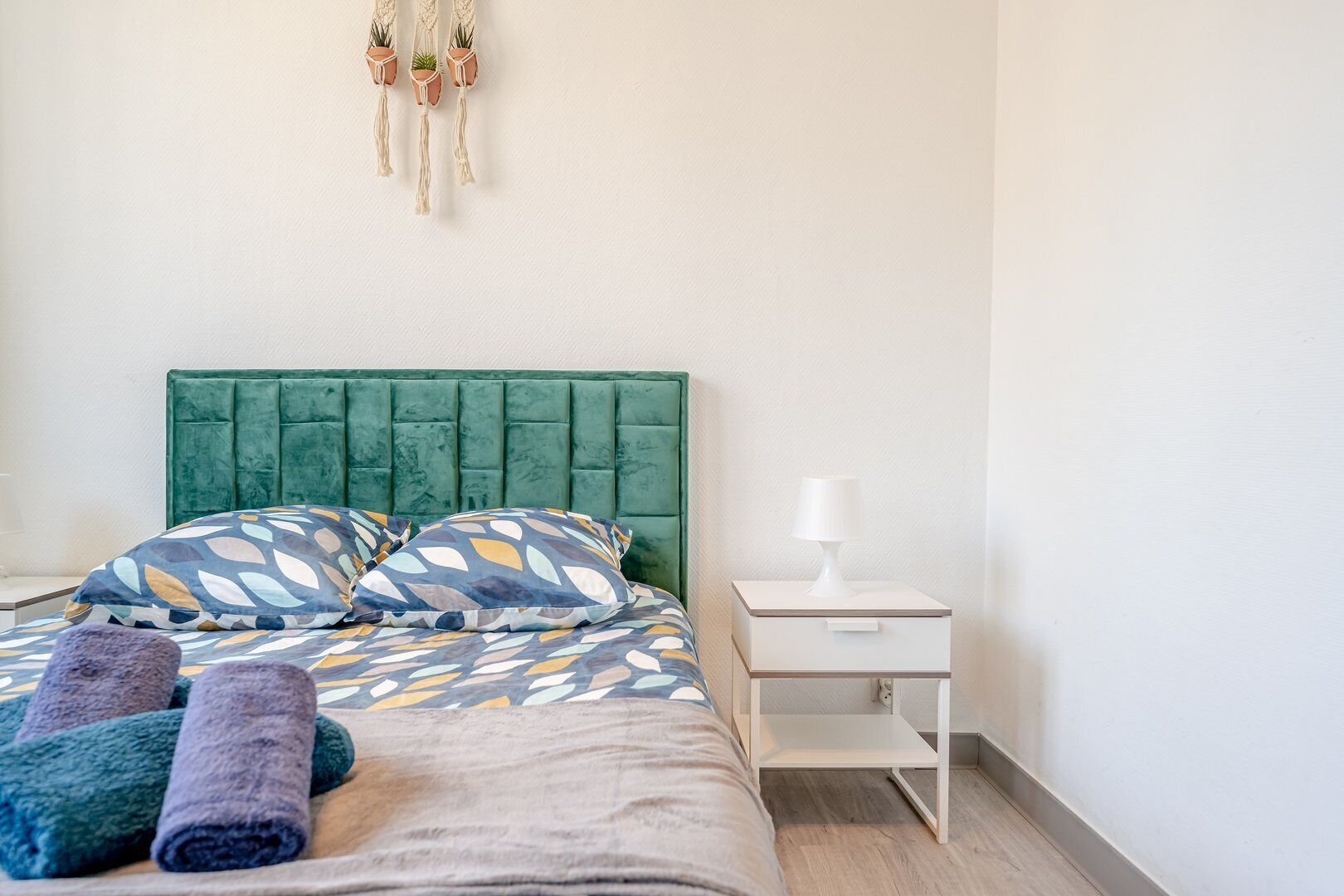 Ein ordentlich bezogenes Bett mit gemusterten Kissen und einem grünen Kopfteil, ein Nachttisch mit einer Lampe sowie eine wandhängende Makramee-Dekoration.