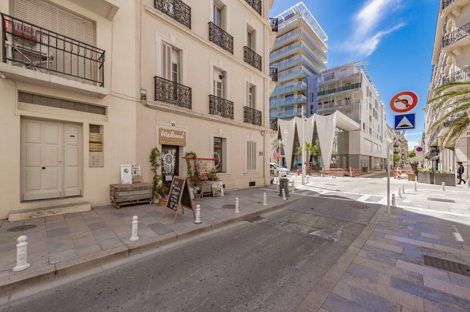 Vista de la calle con un edificio que muestra un negocio en la planta baja, una puerta blanca y un balcón con barandillas negras. El edificio lleva la etiqueta 'Maluna'.