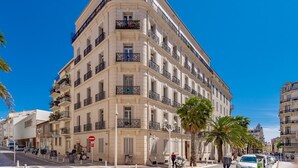A multi-story building with balconies, a street in front, and palm trees.