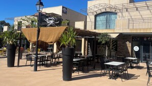 Un espace café en plein air, avec des tables et des chaises, entouré de plantes en pot et d’un drapeau.