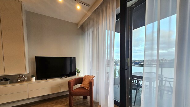 A modern living room with a flat-screen TV on a cabinet, a wooden chair, and a view of a body of water through large windows.