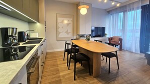 A modern kitchen with a dining area, wooden flooring, and a large window with sheer curtains.