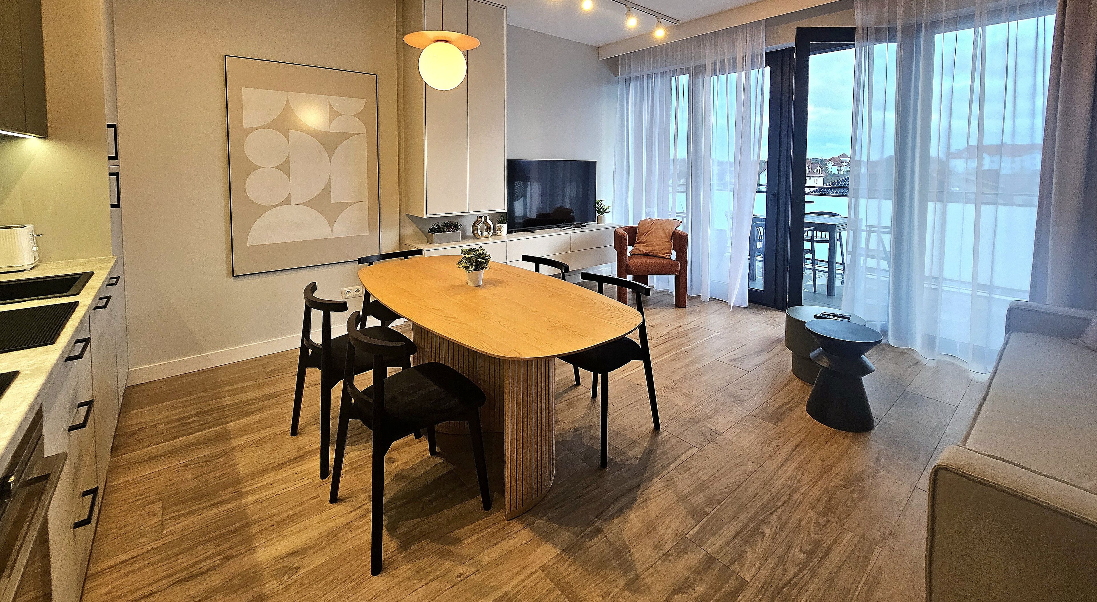 A modern hotel room with a wooden dining table, black chairs, a TV, and a balcony with a view.