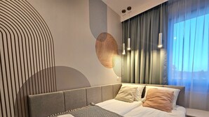 A modern bedroom with a bed, a headboard, a large window with curtains, and a wall with a geometric pattern.