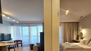 A modern hotel room with a bed, bedside table, and a large window with curtains. There is a dining area with a table and chairs, and a TV mounted on the wall.