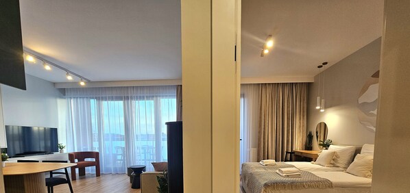 A modern hotel room with a bed, bedside table, and a large window with curtains. There is a dining area with a table and chairs, and a TV mounted on the wall. - Apartament Wiatrem i Piaskiem - 365PAM (Ustronie Morskie)