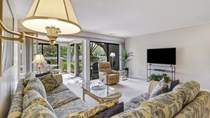 A living room with a sofa set, a glass coffee table, a television on a stand, and a large window with a view of greenery. - Cozy Beach Retreat with Tennis! Short walk to the beach! (Hilton Head Island)