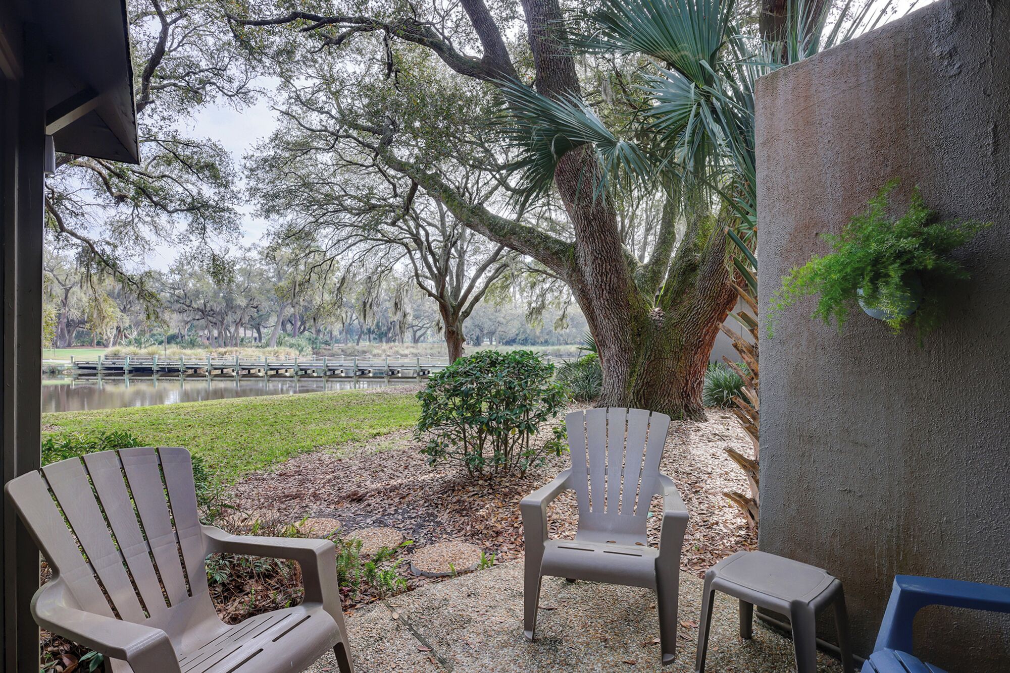 Una zona patio con due poltrone Adirondack e un piccolo sgabello, con vista su un lago e un parco con alberi e un ponte di legno.